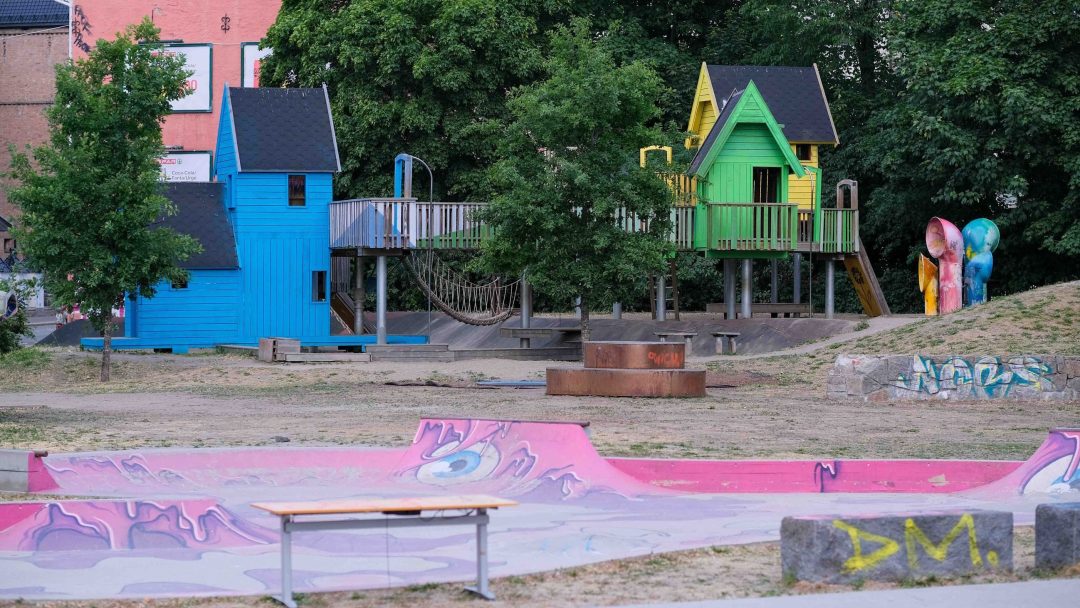 Et pulserende lekeplasser med fargerike lekehus, taubro og sklier venter. I nærheten viser skateparkentusiaster sine ferdigheter blant graffitikunst, alt plassert i en rolig enklave av trær og bygninger.