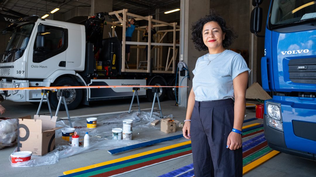 En kvinne står med stolthet ved siden av lastebiler og kunstrekvisita i et yrende verksted.