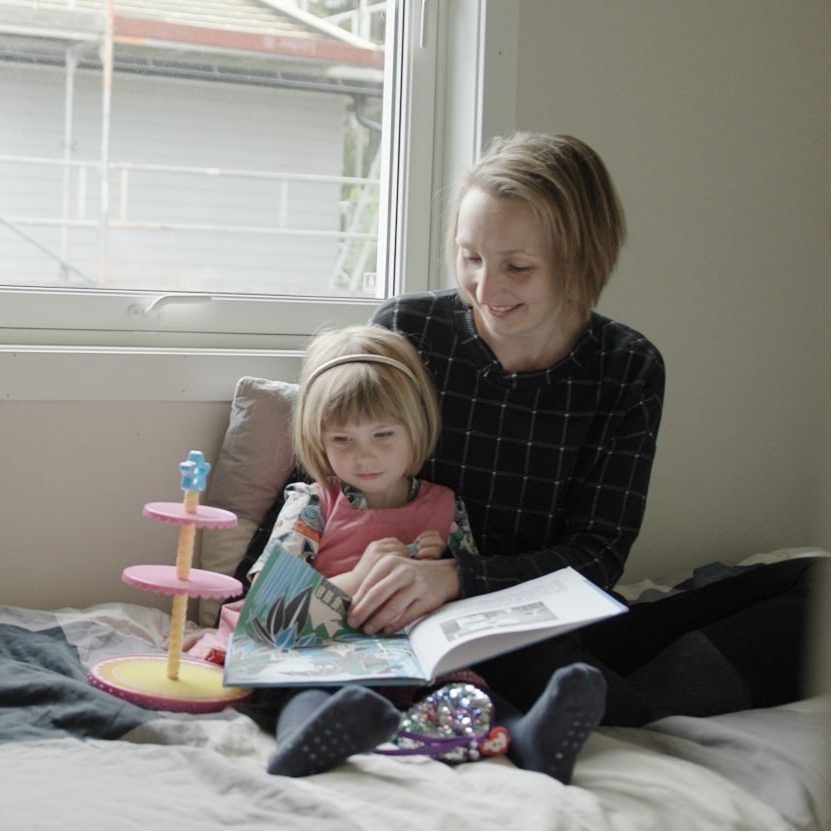 En kvinne og et barn sitter på en seng ved vinduet, badet i myk strøm. Kvinnen holder en åpen bok og fanger barnets blikk. I nærheten hviler et leketøy og sko på sengen, og følger stille med dette rolige øyeblikket.
