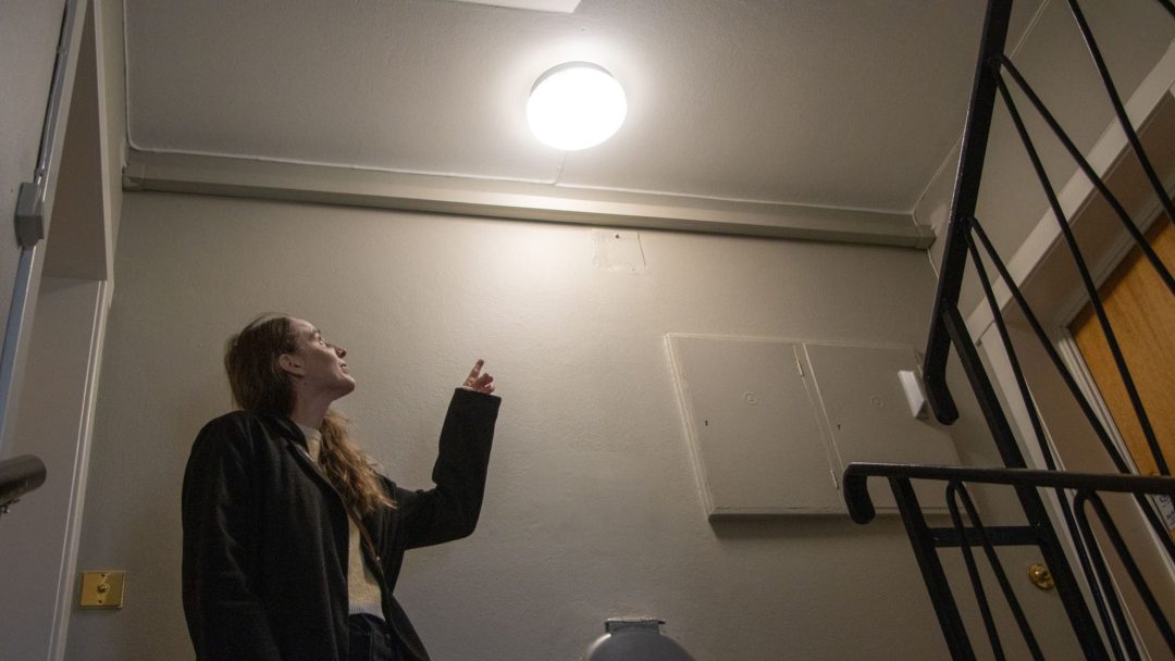 I en gang peker en person på en taklampe, og fremhever potensiell energitiltak. I nærheten er det trapper og en lukket dør.