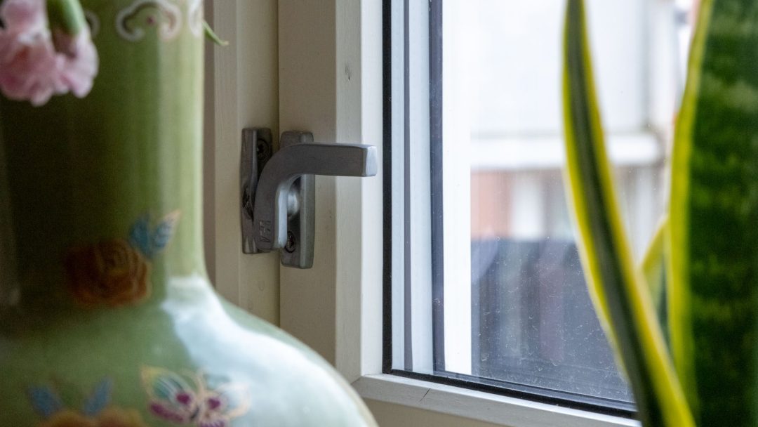Nærbilde av et vindushåndtak ved siden av en dekorativ grønn vase og en høy plante, som fremhever energieffektive fremskritt. Utsikten utenfor er delvis synlig gjennom glasset.