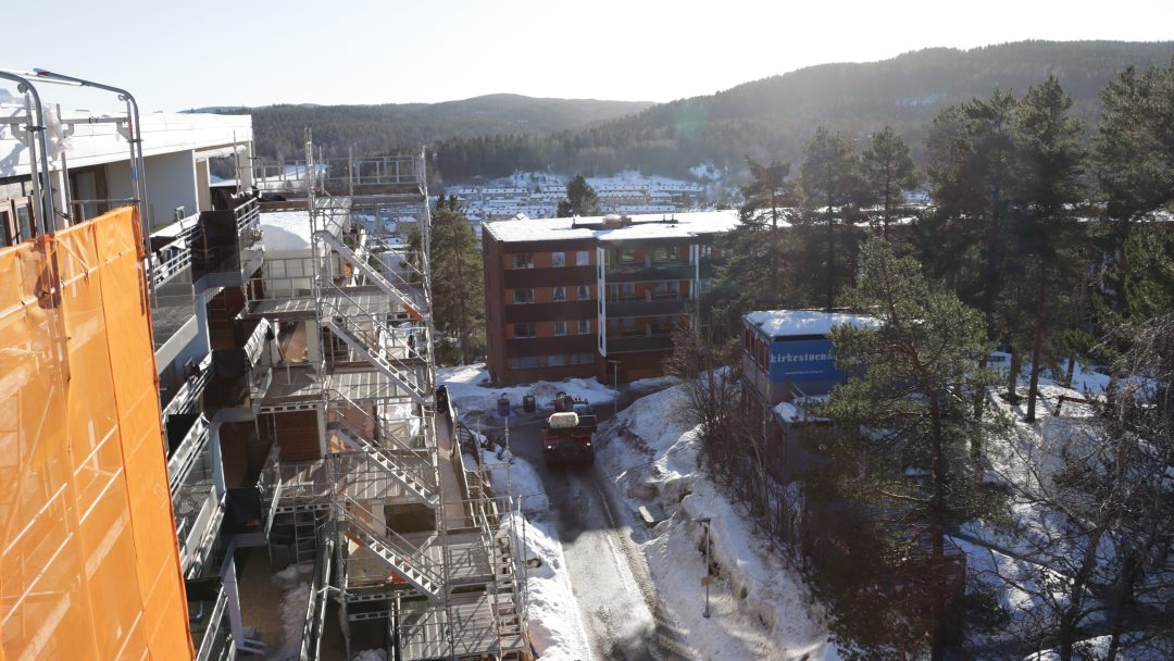 Byggeplassen har stillaser på en bygning, med en lastebil som navigerer en snødekt vei. I bakgrunnen antyder trær og andre bygninger pågående energitiltak under klar himmel, noe som gjenspeiler innsatsen for økt effektivitet.