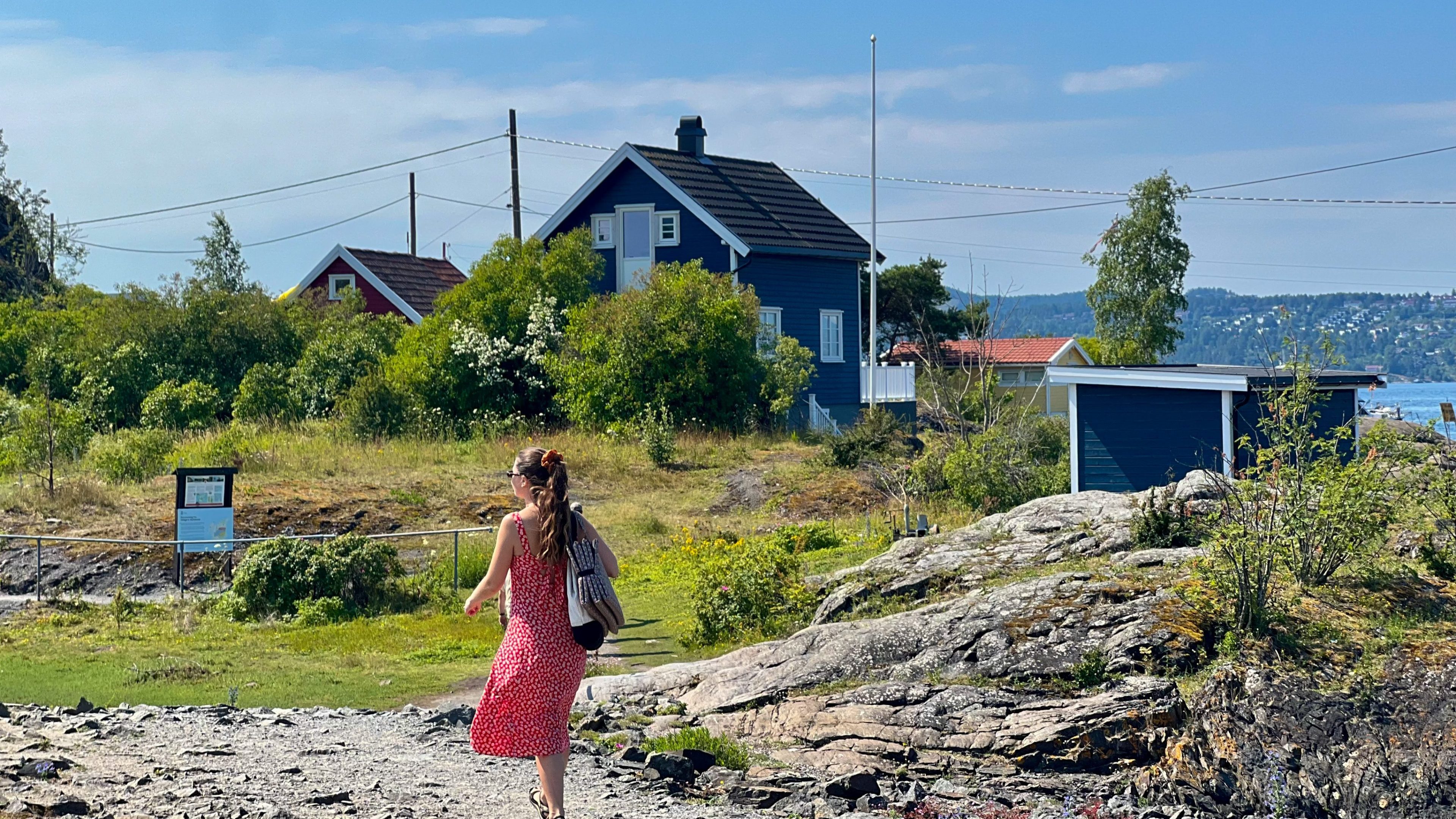 En kvinne i rød kjole navigerer den steinete stien mot et blått hus omsluttet av frodig grønt, som på et øyhopping-eventyr, med en glitrende vannmasse som maler bakgrunnen.