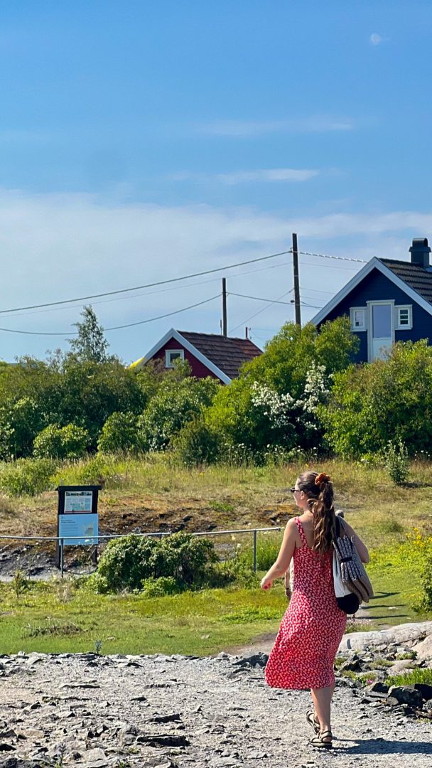En person i rød kjole rusler langs en steinete sti, omgitt av frodige grønne busker og fargerike hus, mens han nyter den rolige skjønnheten til øyhopping under en klarblå himmel.
