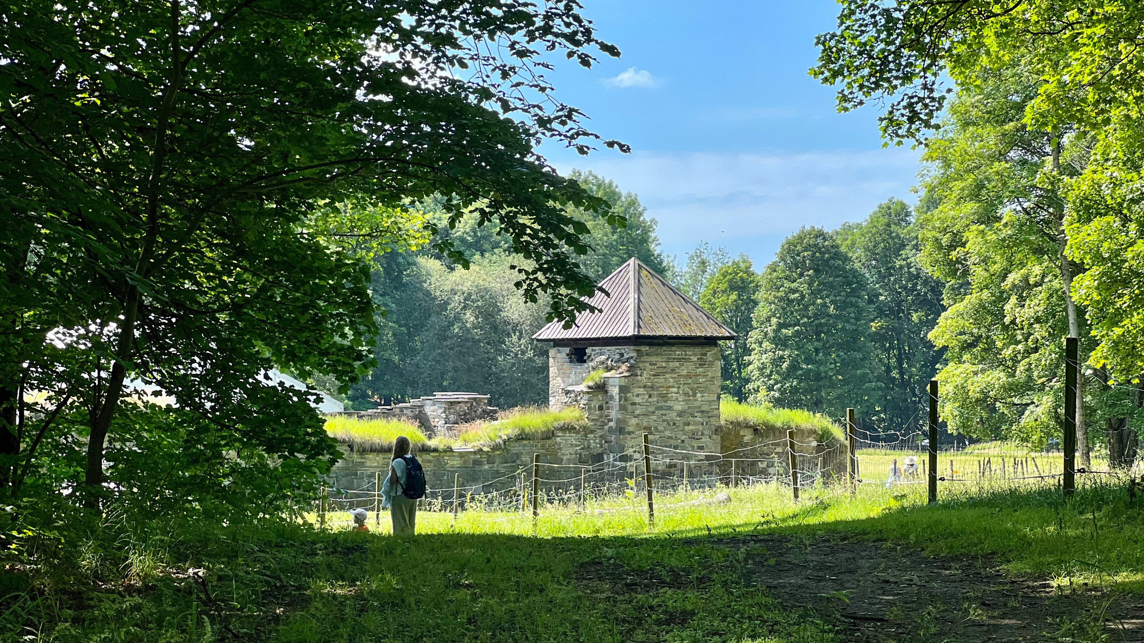 En person står i nærheten av en gammel steinbygning med et konisk tak, omgitt av frodige grøntområder og trær under en klarblå himmel, og mimrer om deres siste øyhoppeeventyr.