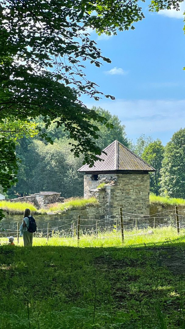 En person med ryggsekk står i nærheten av en liten steinbygning med trekantet tak, omgitt av frodige grøntområder og trær, som om han er klar til å legge ut på et øyhoppeeventyr.