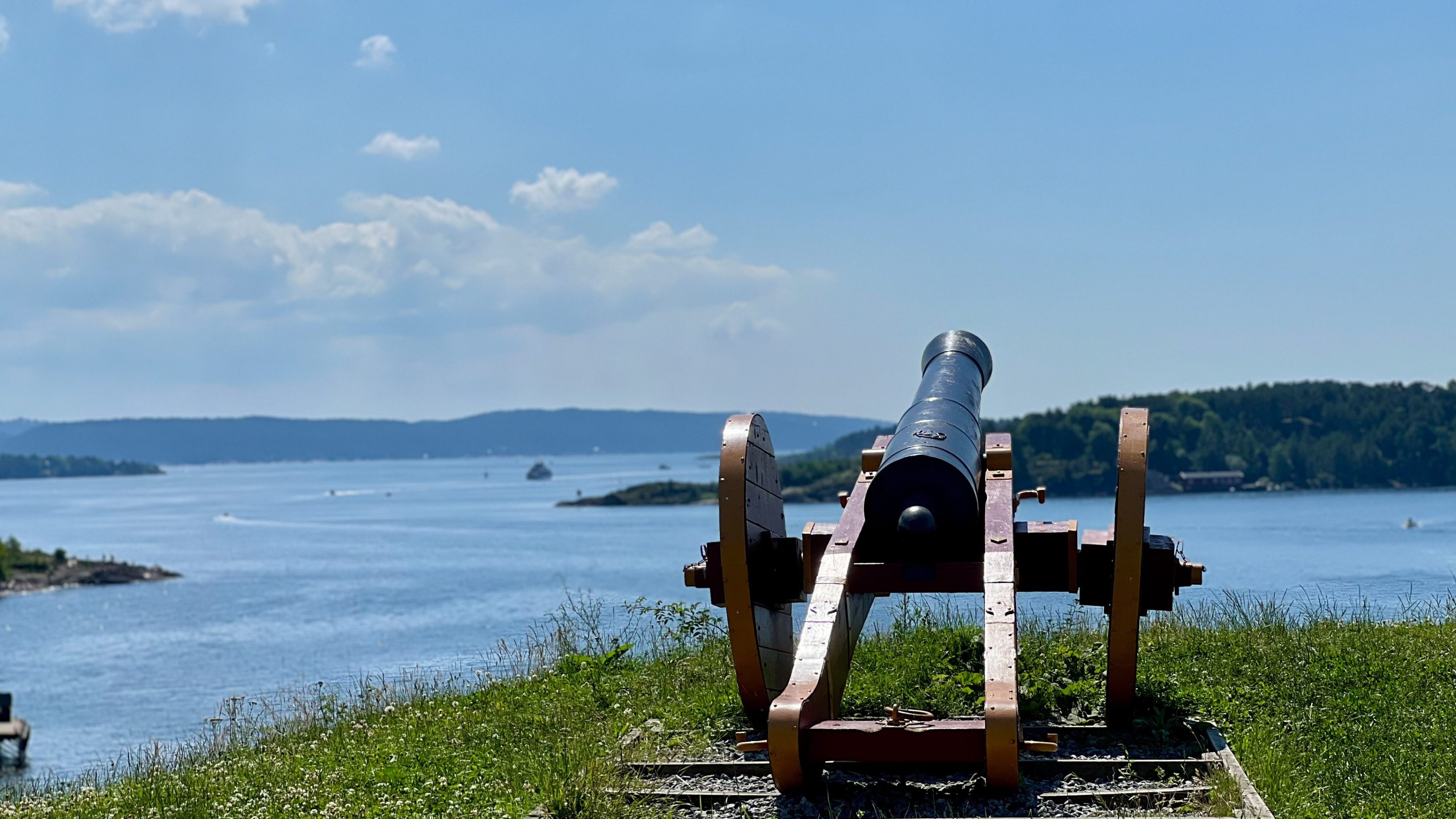 En gammel kanon på en gresskledd bakketopp har utsikt over en vannmasse med fjerne øyer, perfekt for øyhopping-eventyr, under en klarblå himmel.