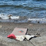 En rolig strandscene utspiller seg med en matte lagt på småstein, omgitt av spredte klær og sandaler. En bok hviler innbydende på matten, mens bølger forsiktig slår mot kysten – perfekt for å slappe av etter øyhopping-eventyr.