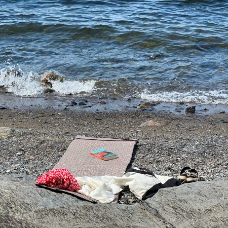 En rolig strandscene utspiller seg med en matte lagt på småstein, omgitt av spredte klær og sandaler. En bok hviler innbydende på matten, mens bølger forsiktig slår mot kysten – perfekt for å slappe av etter øyhopping-eventyr.