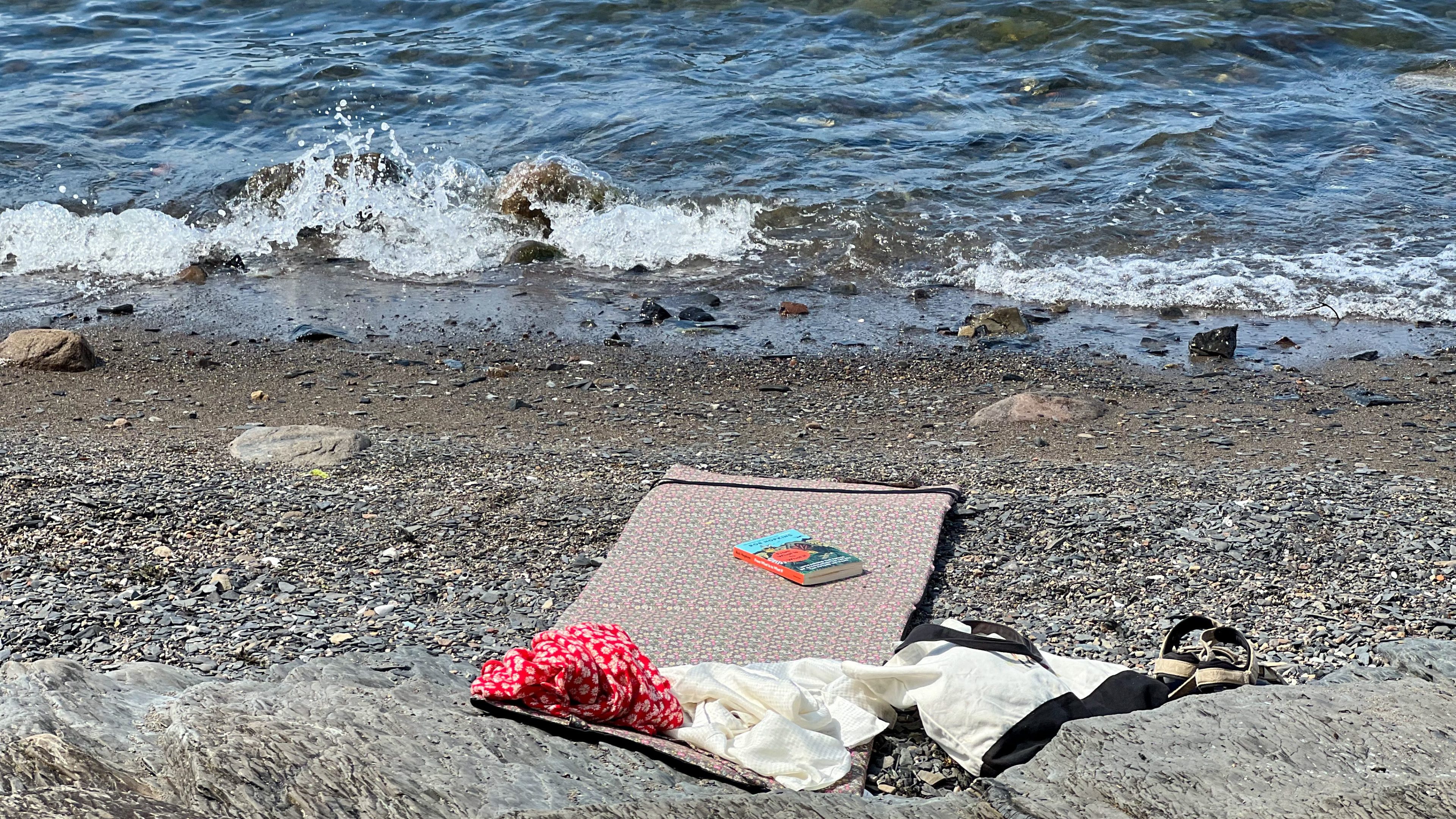 En rolig strandscene utspiller seg med en matte på småstein, en bok plassert på toppen, omgitt av en rød mønstret klut, hvit veske og svarte sandaler. Mens bølger forsiktig slår mot kysten, går tankene til øyhoppende eventyr utenfor horisonten.