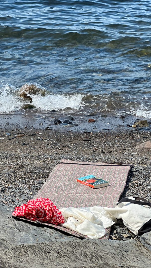 En rolig strandscene har en mønstret matte på småstein, et rødt og hvitt plagg, et hvitt plagg og en bok, som inviterer til avslapning etter øyhopping-eventyr. I bakgrunnen slår bølgene forsiktig mot kysten.