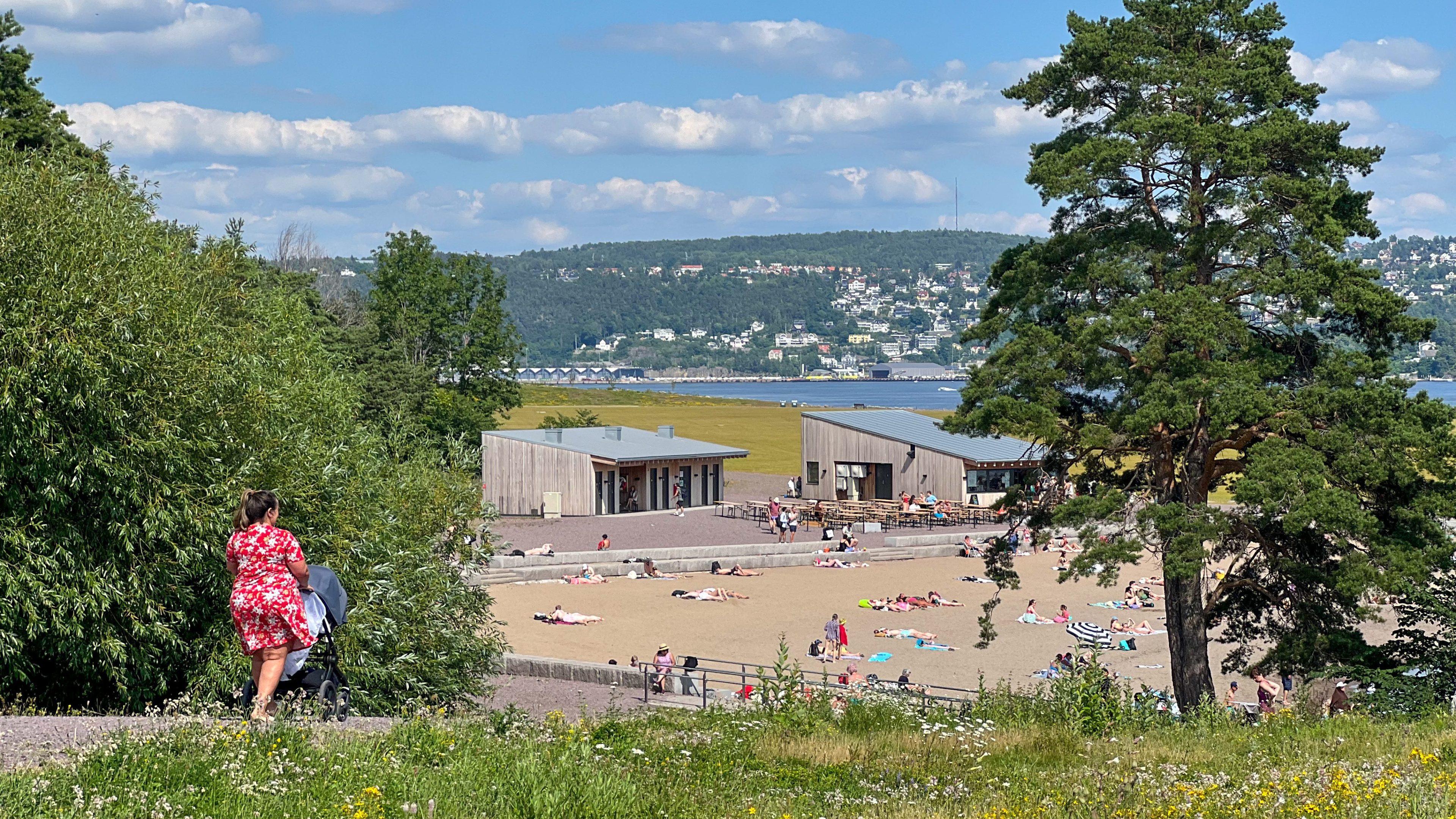 Folk slapper av på en sandstrand nær et gressområde og moderne bygninger, med grønne åser og en vannmasse i bakgrunnen, perfekt for å planlegge ditt neste øyhoppeeventyr.