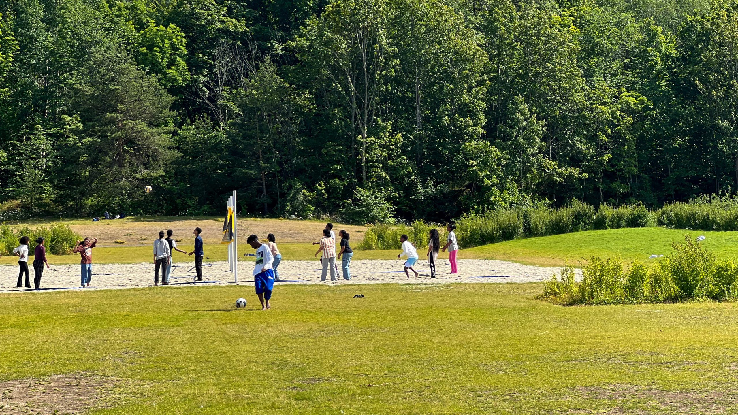 En gruppe mennesker rusler gjennom en gresskledd park, med trær som svaier i bakgrunnen. Midt i disse rolige omgivelsene nærmer en person seg blått en fotball, og legemliggjør ånden av øyhopping med hvert livlige skritt mot den.
