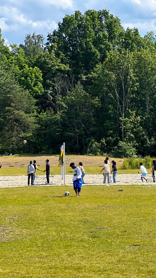 Midt i frodige grøntområder liker folk volleyball på en sandbane mens andre ser på. I forgrunnen hviler en fotball på gresset, kanskje venter på en pause fra spillet for å planlegge sitt neste øyhoppeeventyr.