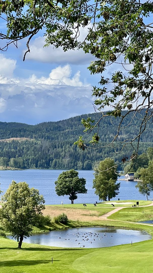 Vakker utsikt over en golfbane med et lite tjern, trær og en innsjø i bakgrunnen under en delvis overskyet himmel, alt sammen med pittoreske sykkelstier for sykkelentusiaster.