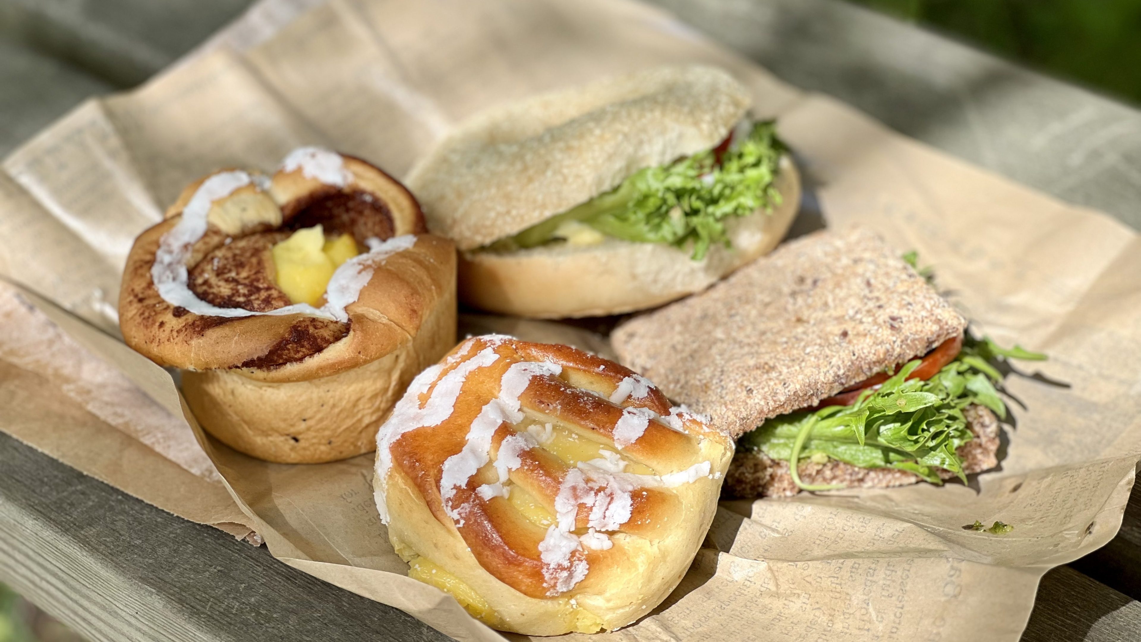 Fire bakverk og smørbrød kunstferdig arrangert på en treflate, med en søt bolle, en eggesmørbrød og en rugbrødssmørbrød – perfekt drivstoff for ditt neste eventyr langs sykkelstien.