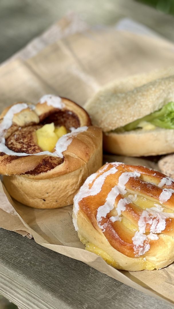 Tre bakverk hviler på et papir som dekker en treflate: et rundt bakverk med glasur, et annet fylt med vaniljesaus og et smørbrød med salat – et herlig stopp for syklister som utforsker sykkelstien.