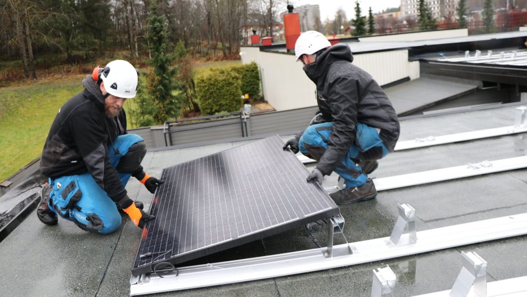 To arbeidere i verneutstyr installerer flittig et solcellepanel på et hustak, omgitt av grøntområder og bygninger, som en del av lokale energitiltak-tiltak.