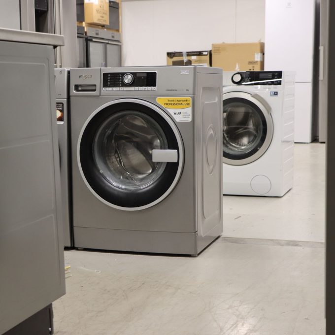 To vaskemaskiner er utstilt i et utstillingsrom, en sølv og en hvit, begge med digitalt kontrollpanel. Energimerkingen er tydelig vist, og indikerer deres effektivitet. Bokser og hyller er synlige i bakgrunnen, og fremhever et bredt utvalg av elektriske produkter tilgjengelig.