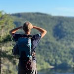 En person med ryggsekk nyter en topptur, stirrer på en skogkledd dal fra et utsiktspunkt, omgitt av trær under en klarblå himmel.