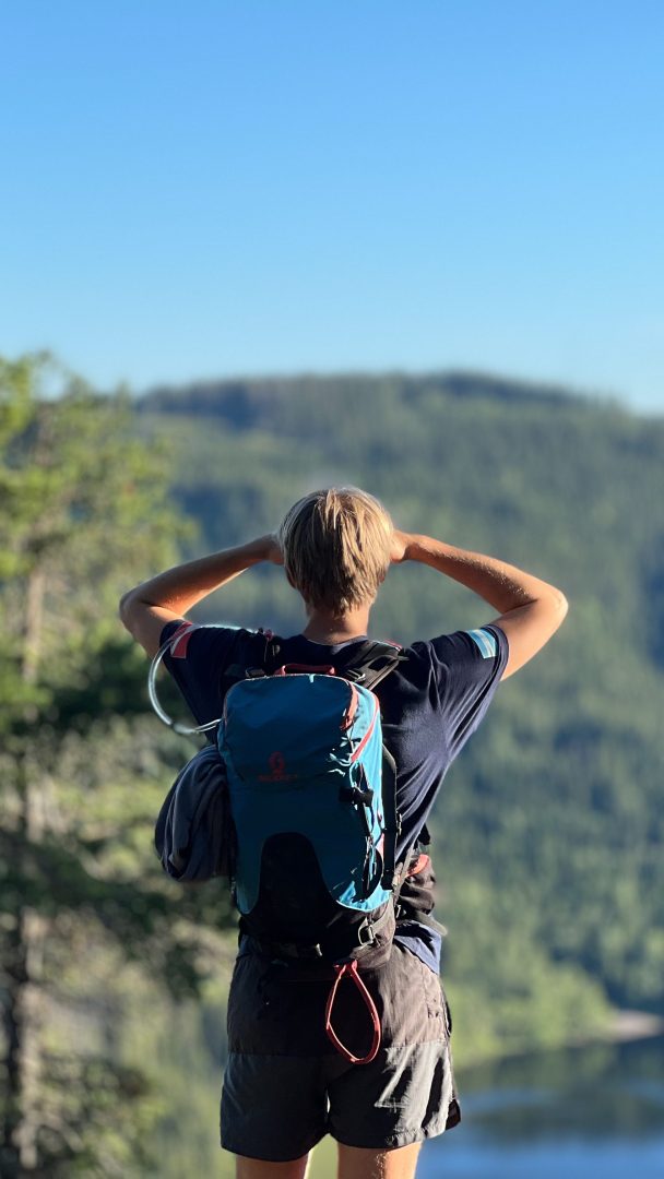 En ensom eventyrer med ryggsekk tar en pause på toppen av bakken, og omfavner toppturs ånd mens de ser på et fjernt skoglandskap under en klarblå himmel.