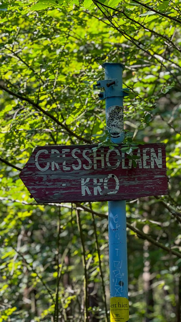 Et rødt treskilt med «GRESSHOLMEN KRO» i hvite bokstaver står stolt på en blå stang og inviterer de på øyhopping-eventyr, omgitt av frodig grønt løvverk.