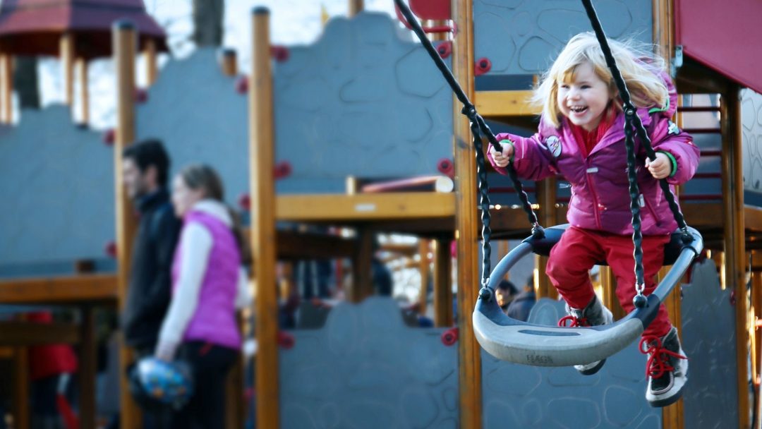 Et barn i rosa frakk smiler gledesfylt på en huske på lekeplassene, mens to voksne ser fornøyde på i bakgrunnen.