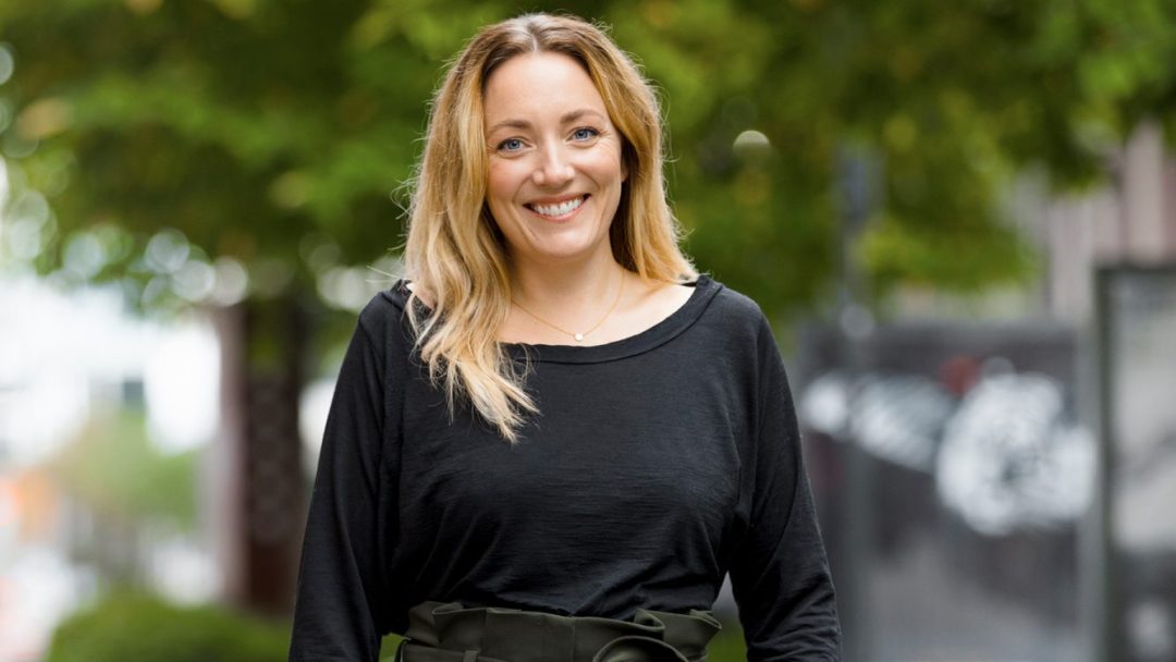 En smilende person med langt hår, iført en svart topp, står utendørs blant frodige trær, mens det milde skjæret fra overvann skaper et rolig bakteppe.