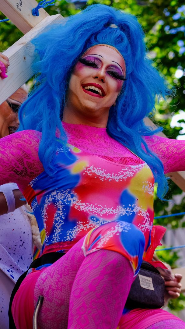 En person i livlig sminke og et fargerikt antrekk, med blå parykk og rosa kjole, utstråler stolthet mens de smiler i en utendørs setting.