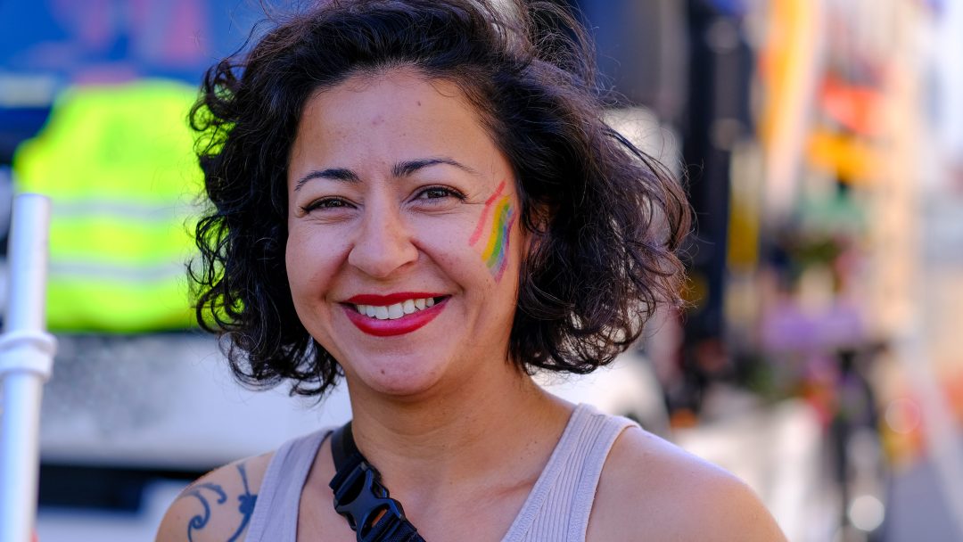 En person med kort krøllete hår og en regnbue malt på kinnet, smilende utendørs, utstråler stolthetens ånd.