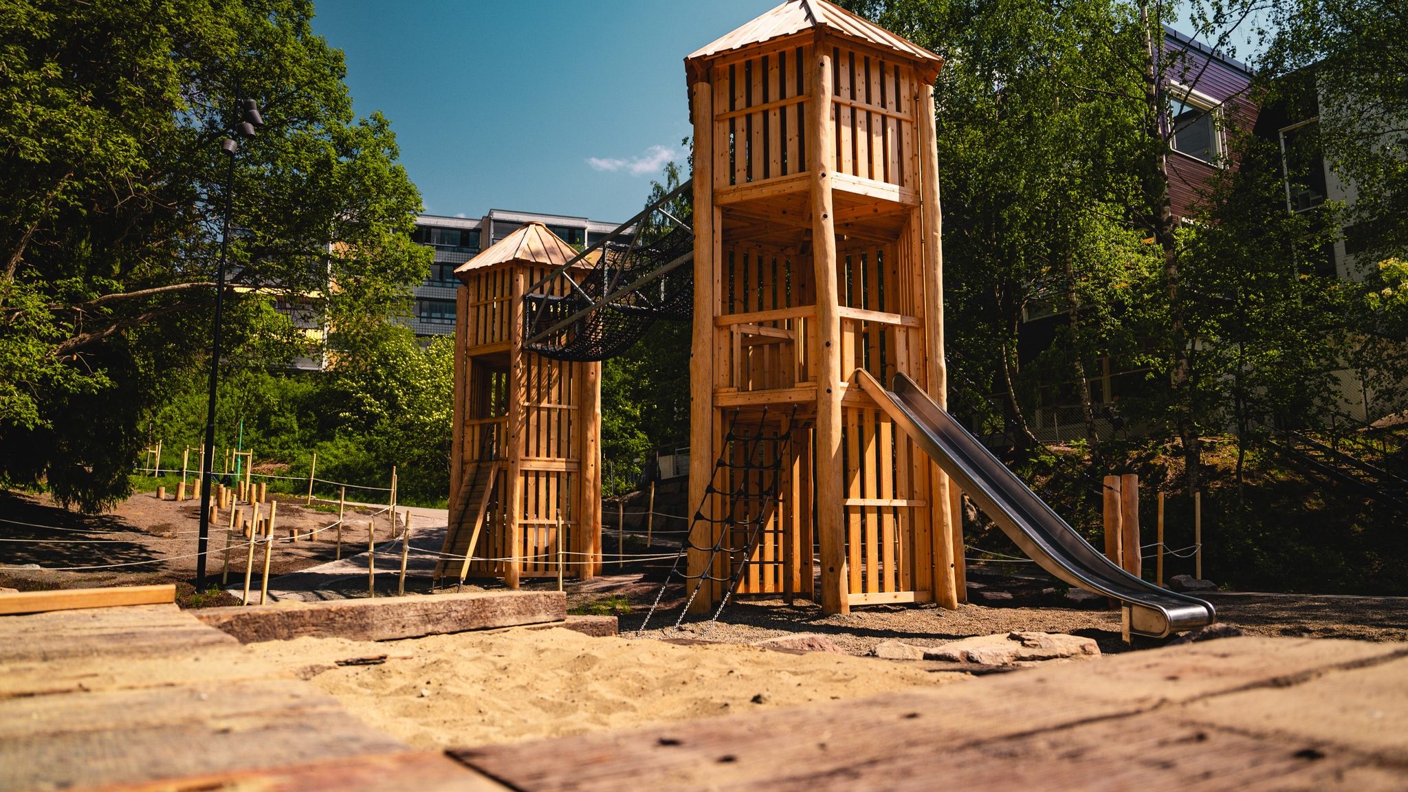 En sjarmerende lekeplasser i tre har to tårn forbundet med en bro, komplett med spennende sklier og utfordrende klatreområder. Omsluttet av trær og bygninger, soler dette lekne tilfluktsstedet under en solfylt himmel, og byr på uendelig moro for barn i livlige omgivelser.