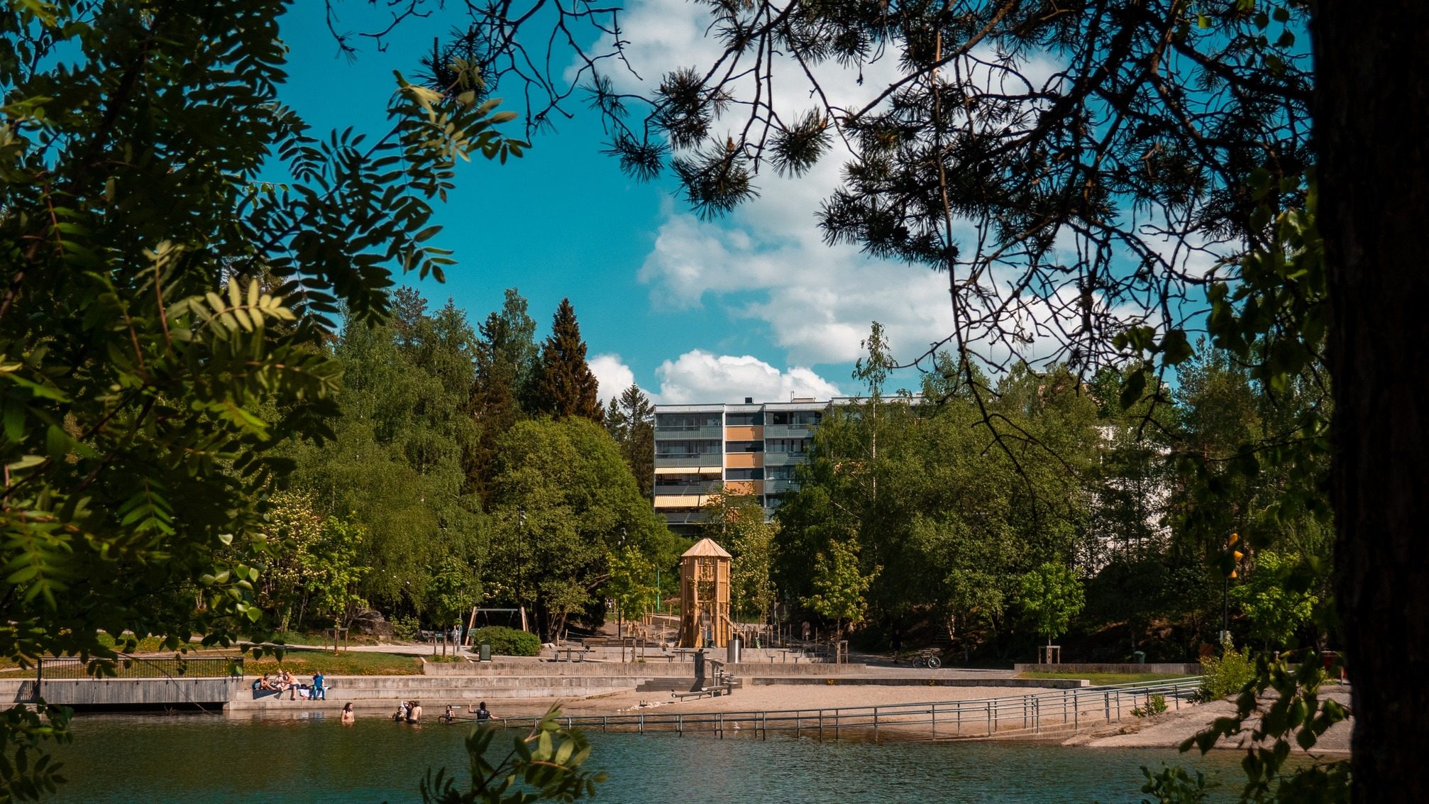 En parkscene ved innsjøen har en moderne leilighetsbygning omgitt av trær. I nærheten slapper folk av på en brygge og svømmer under en klarblå himmel, mens barna koser seg på lekeplassene i nærheten.