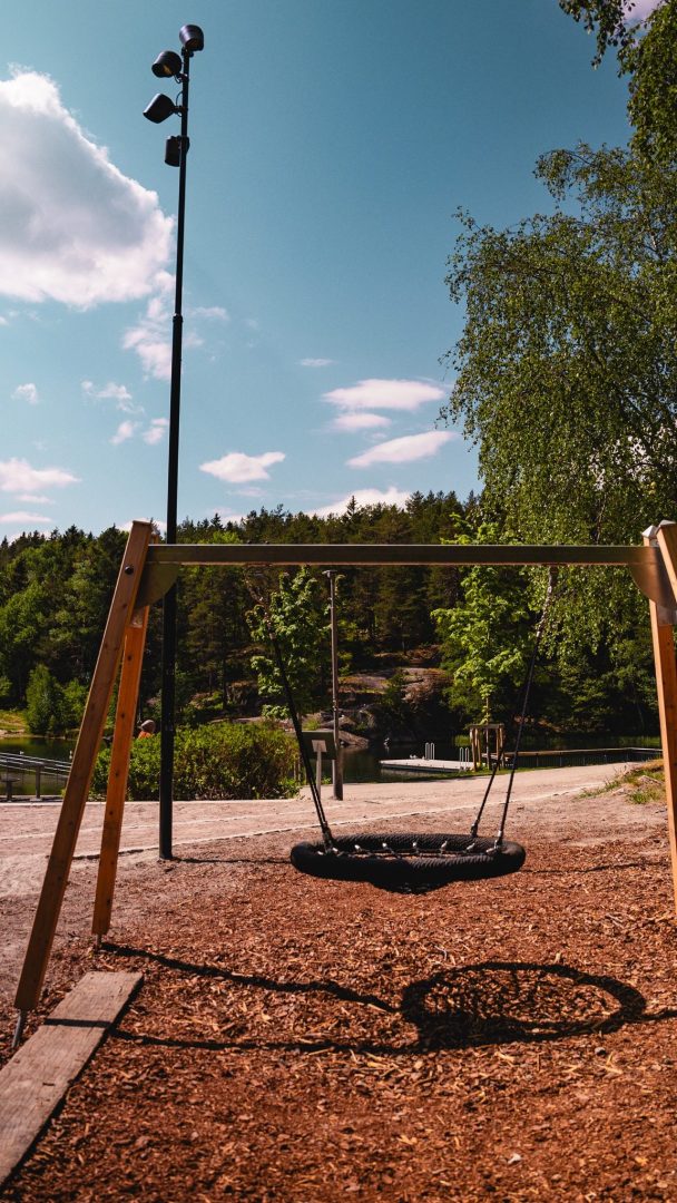 En enkelt rund huske henger fra en treramme i et pulserende lekeplasser. Rundt det er trær, en gatelykt og en blå himmel oversådd med skyer, og skaper det perfekte bakteppet for bekymringsløs lek.