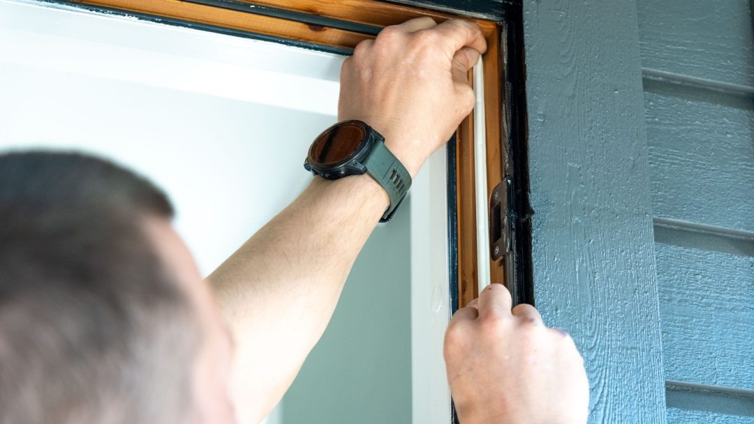 En person med klokke justerer en treramme på en dør eller et vindu mot en mørk yttervegg, og integrerer energieffektive tiltak som en del av energitiltak.