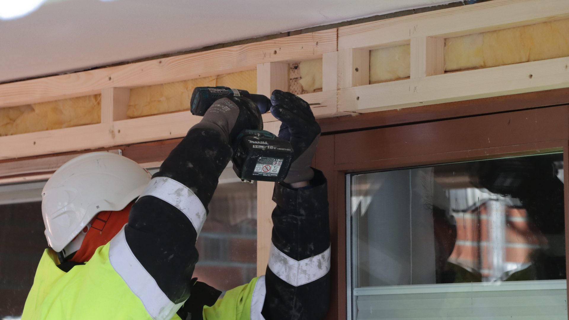 En bygningsarbeider i vernehjelm og synlig jakke fokuserer på energitiltak, ved hjelp av en boremaskin for å sikre treinnramming over et vindu.