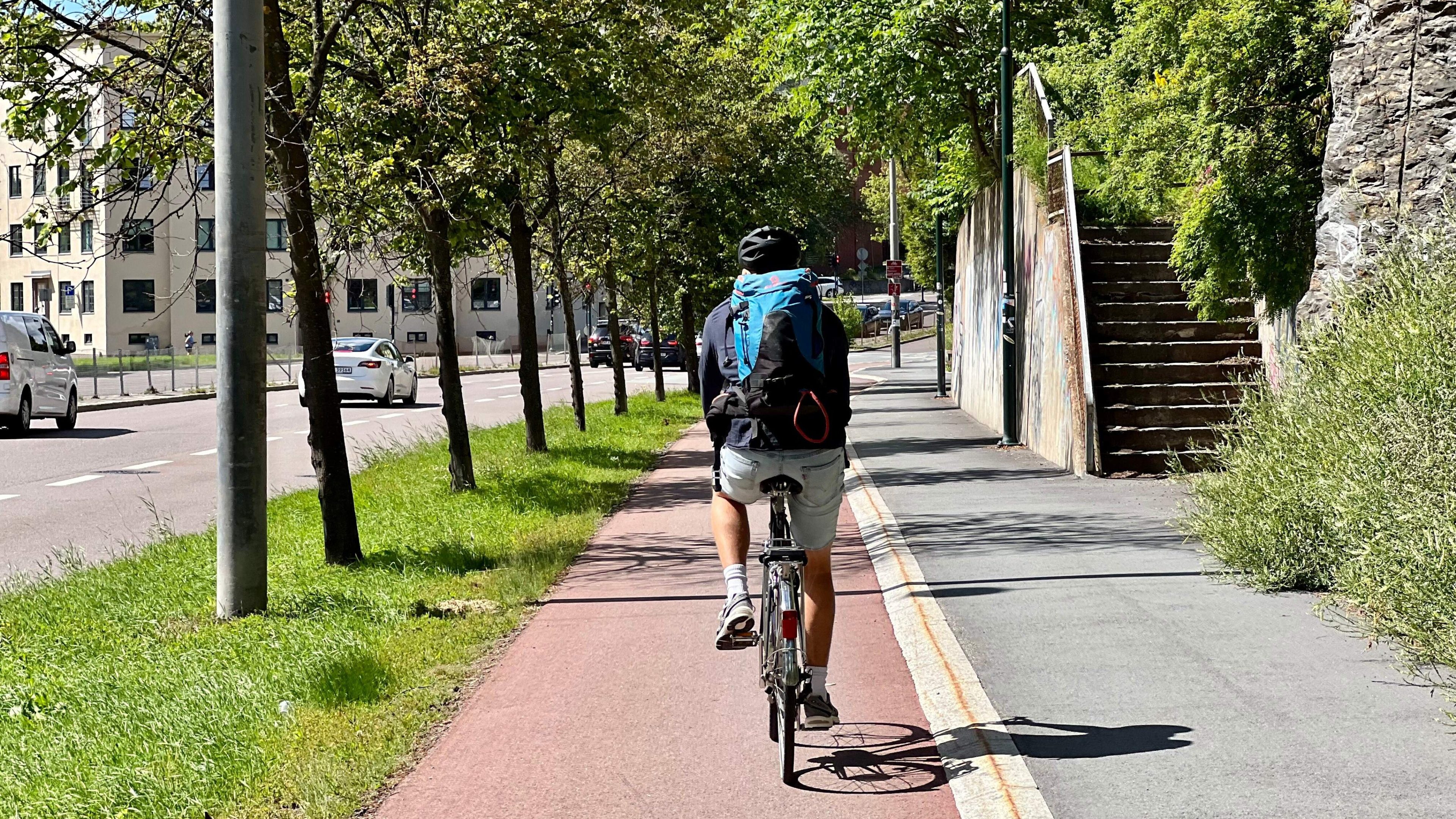 En person tråkker langs en asfaltert sykkelstier ved siden av en vei, flankert av høye trær og en trapp i nærheten, alt under klar himmel.