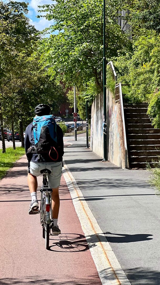 En person sykler langs en sykkelsti, i skyggen av trær, ved siden av en gangvei med trapper.