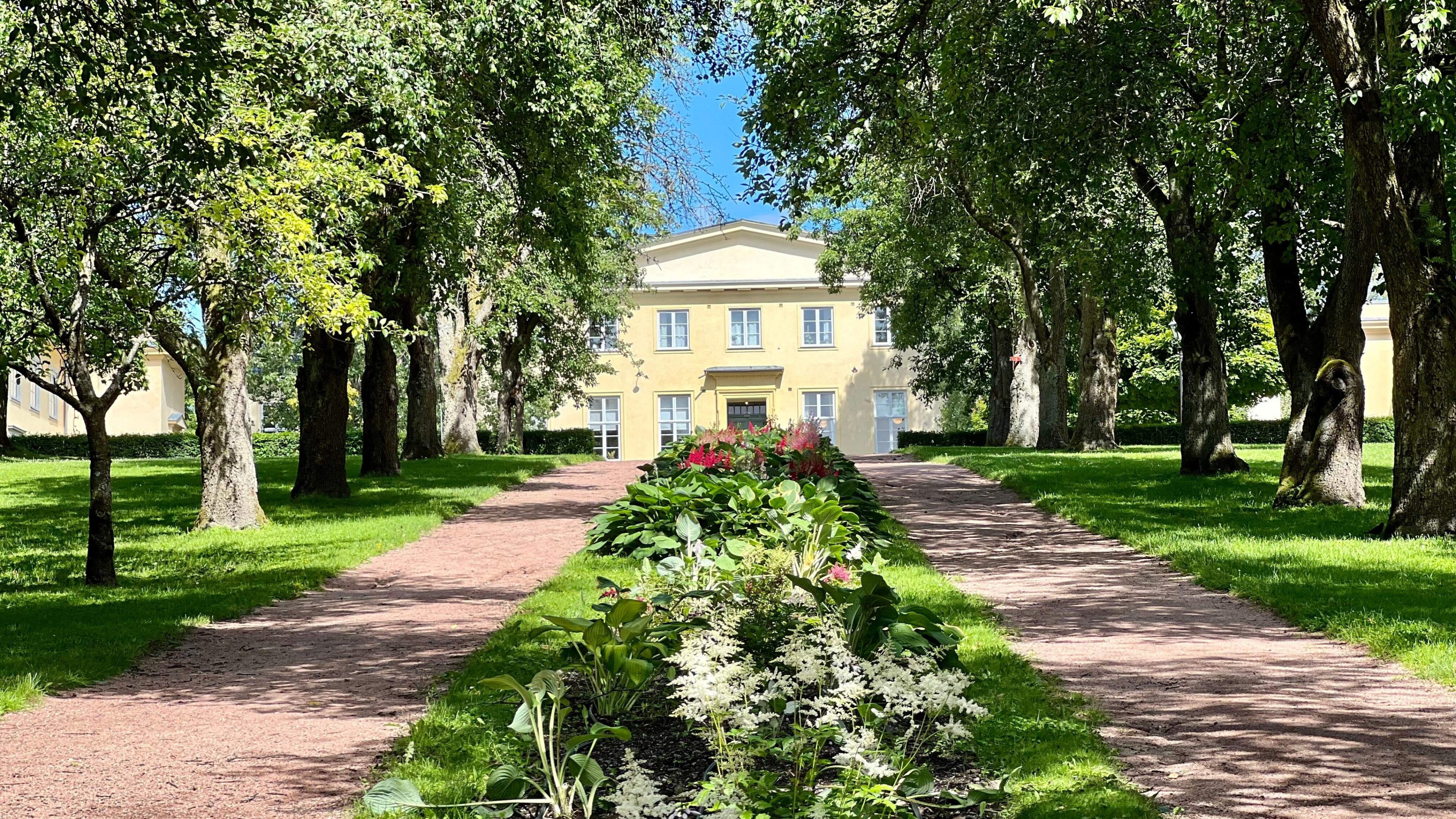 Gul bygning innrammet av trær og en symmetrisk hage med et sentralt blomsterbed i forgrunnen. To grusstier går parallelt på hver side, som minner om sjarmerende sykkelstier som inviterer syklister til å snirkle seg gjennom det rolige landskapet.