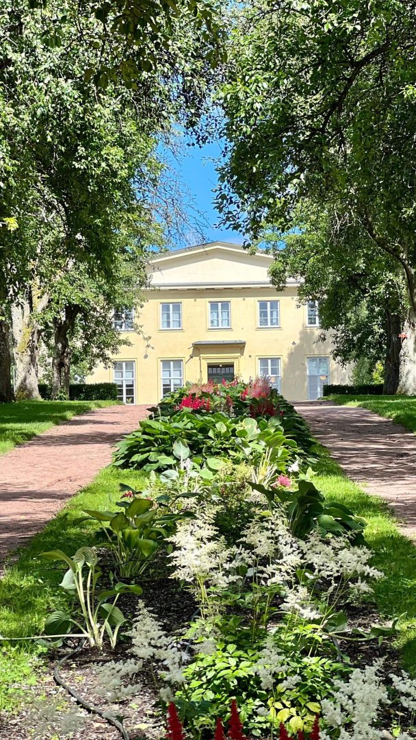 En sti, som ligner rolig sykkelstier, fører til en to-etasjers gul bygning omgitt av frodige grøntområder og levende blomster under en klarblå himmel.