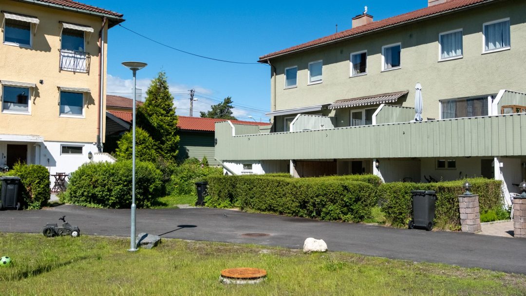 Et boligområde med to livlige bygårder, en gul og en grønn, ligger under en klarblå himmel. Et gresskledd område med en lyktestolpe, søppelkasser og et lite lekekjøretøy som hviler på fortauet fullfører scenen. Nabolaget klarer ettertenksomt overvann å holde alt uberørt.