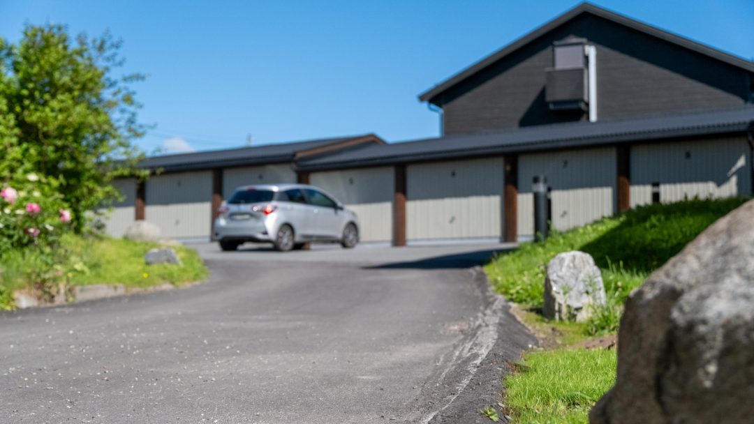 En sølvfarget bil kjører jevnt opp den asfalterte oppkjørselen mot en rad med garasjeporter under klarblå himmel, med grønt på siden som glinser etter et overvann.