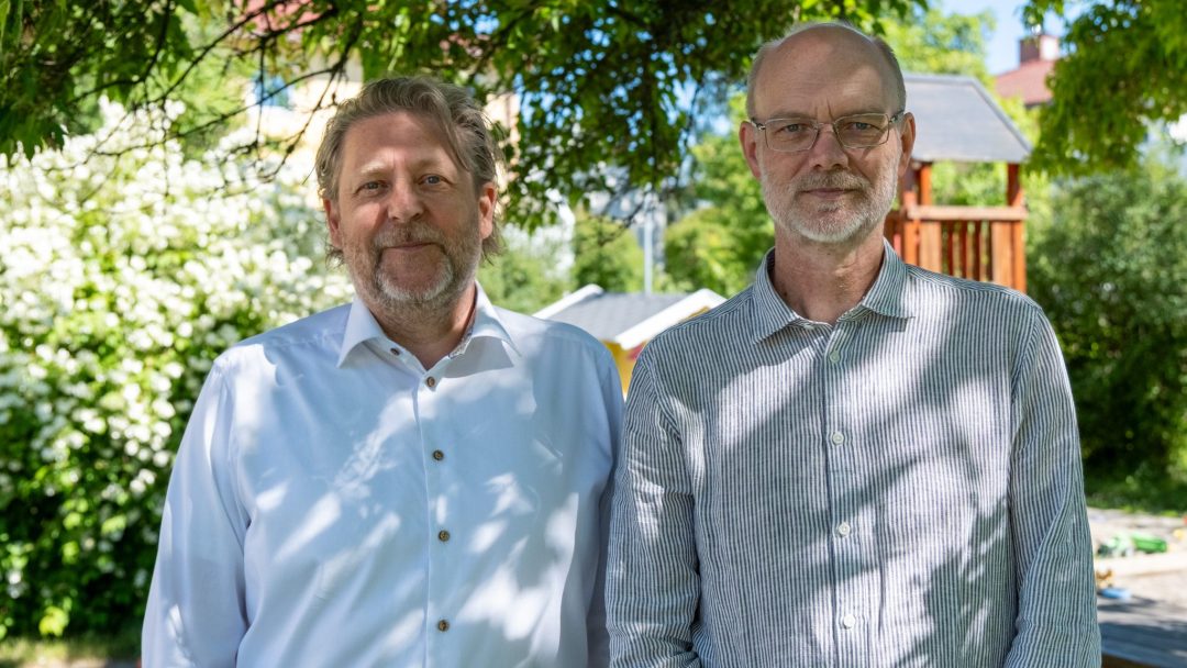 To menn står utendørs under et tre, med lekestativ og frodig grønt i bakgrunnen. Begge har på seg skjorter med krage – den ene hvit, den andre stripete – mens de nyter en dag upåvirket av overvann.