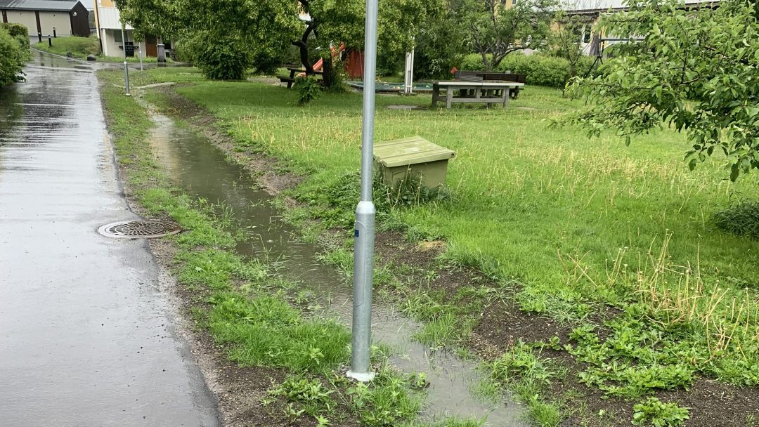 En smal, vannfylt sti med vannpytter går langs et gressområde. Overvann reflekterer lys som en gatelykt og benk er synlig i nærheten av trær og hus. Fortauet er vått av regn.