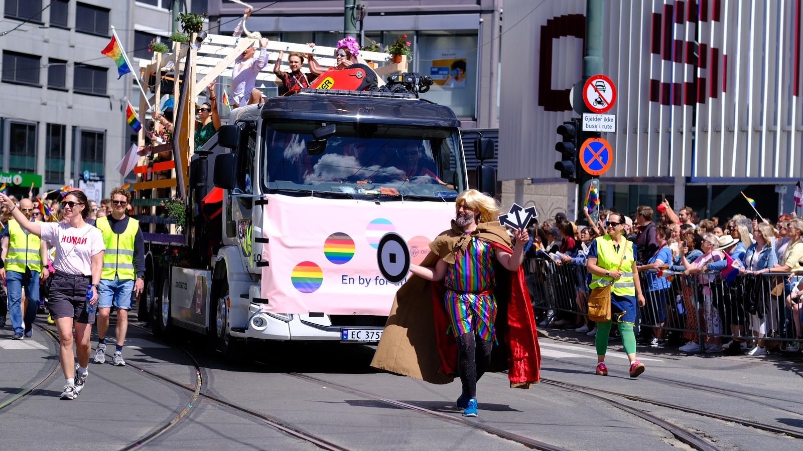 En pride parade flyter utsmykket med regnbueflagg beveger seg nedover gaten, omgitt av mennesker i fargerike kostymer og entusiastiske tilskuere, som feirer under en levende offentlig begivenhet.