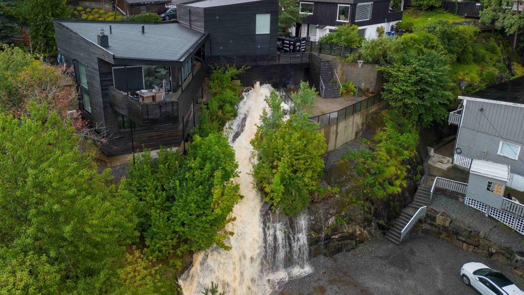 Luftfoto av en foss som fosser ned en åsside, med moderne hus plassert ved siden av og omgitt av frodig grønt, og skaper en pittoresk scene som minner om et overvannslandskap.