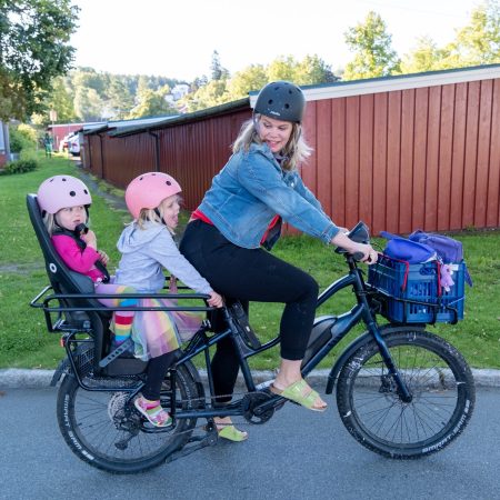 En kvinne kjører en lastesykkel med to barn med hjelmer bak. Sykkelens kurv er fylt med gjenstander mens de kjører langs en vei omgitt av grøntområder og et tregjerde i bakgrunnen.