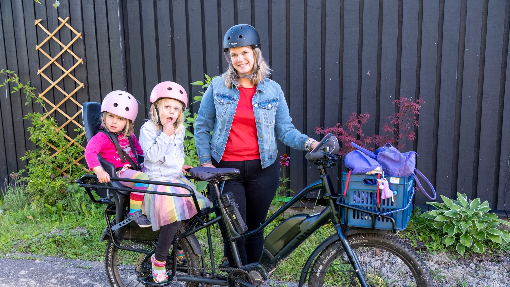 En kvinne i dongerijakke og hjelm står selvsikkert med lastesykkelen sin. To barn, også iført hjelm, sitter i seter festet til sykkelen, den ene gumler på en matbit. En kurv fylt med poser er sikret på baksiden av den solide lastesykkelen.