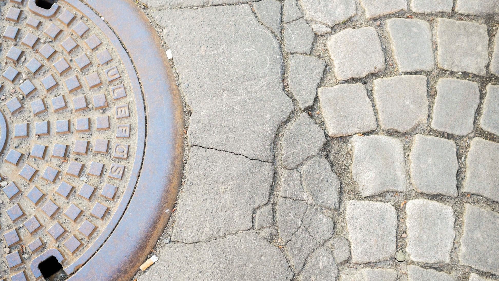 Et nærbilde av et kumlokk satt inn i et sprukket fortau, sammen med en seksjon av brostein, fremkaller sammenstillingen av urban grus og naturens preg, som minner om trær langs bygatene.