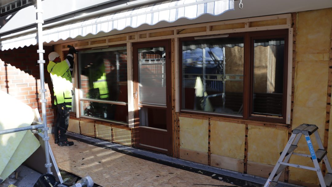 En bygningsarbeider i en gul sikkerhetsvest omhyggelig etterisolere bygningens yttervegg. Veggen er delvis strippet, og avslører lag med gul isolasjon. En stige står på høyre side, klar for neste fase med å befeste strukturens termiske skjold.