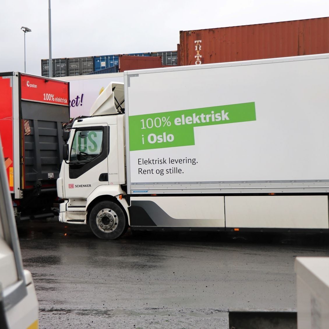 En el-lastebil i Oslo har en grønn og hvit annonse som proklamerer 100% elektrisk i Oslo. Elektrisk levering. Rent og stille. Den er parkert ved siden av en rød lastebil på den våte veien.